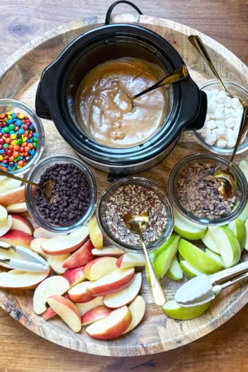 Overhead view of a caramel apple board with a mini crockpot of caramel, apple slices, and various toppings.