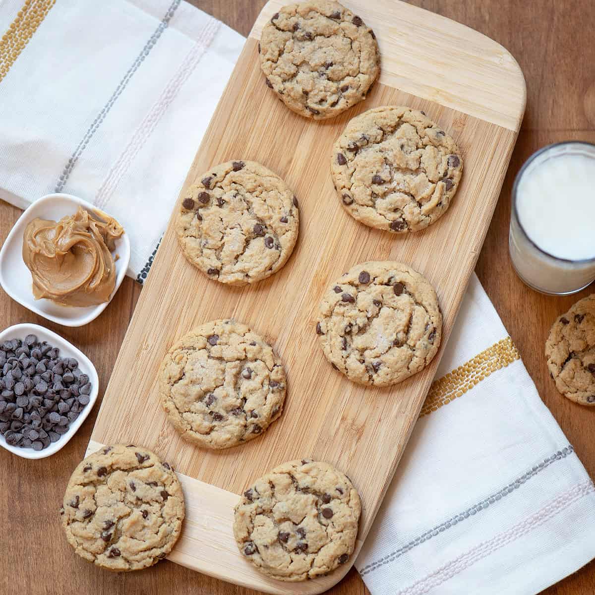 Overhead close-up of peanut butter chocolate cookies on a wooden board with a glass of milk and extra cookies in the background.