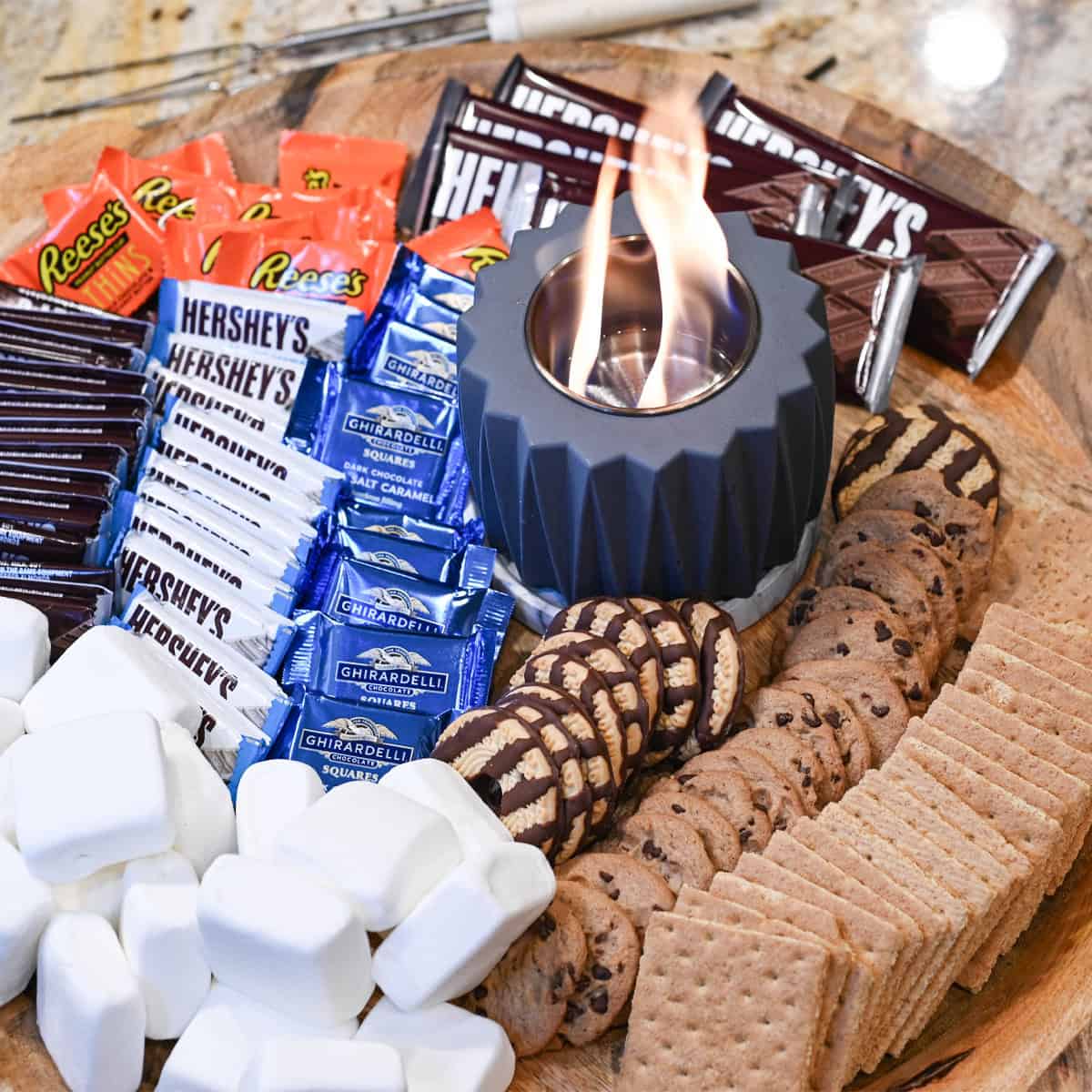 Indoor s’mores board with a tabletop firepit surrounded by marshmallows, chocolate, graham crackers, and assorted toppings.
