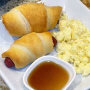 Crescent roll-wrapped sausage links served warm with a side of syrup on a white plate.