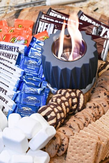 Overhead view of a s’mores board with chocolate, marshmallows, cookies, and a lit tabletop fire pit.
