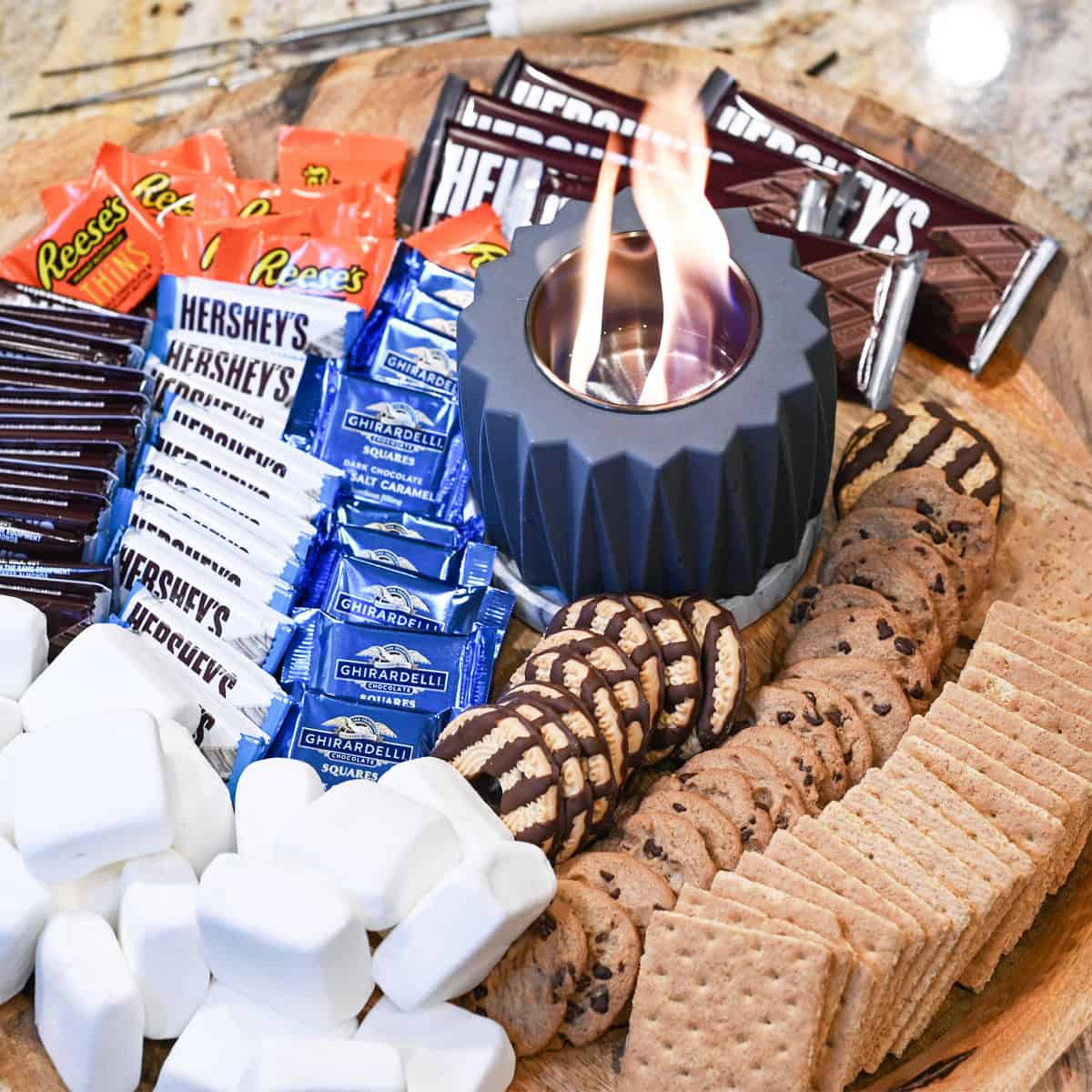 Overhead view of a s’mores board with chocolate, marshmallows, cookies, and a lit tabletop fire pit.