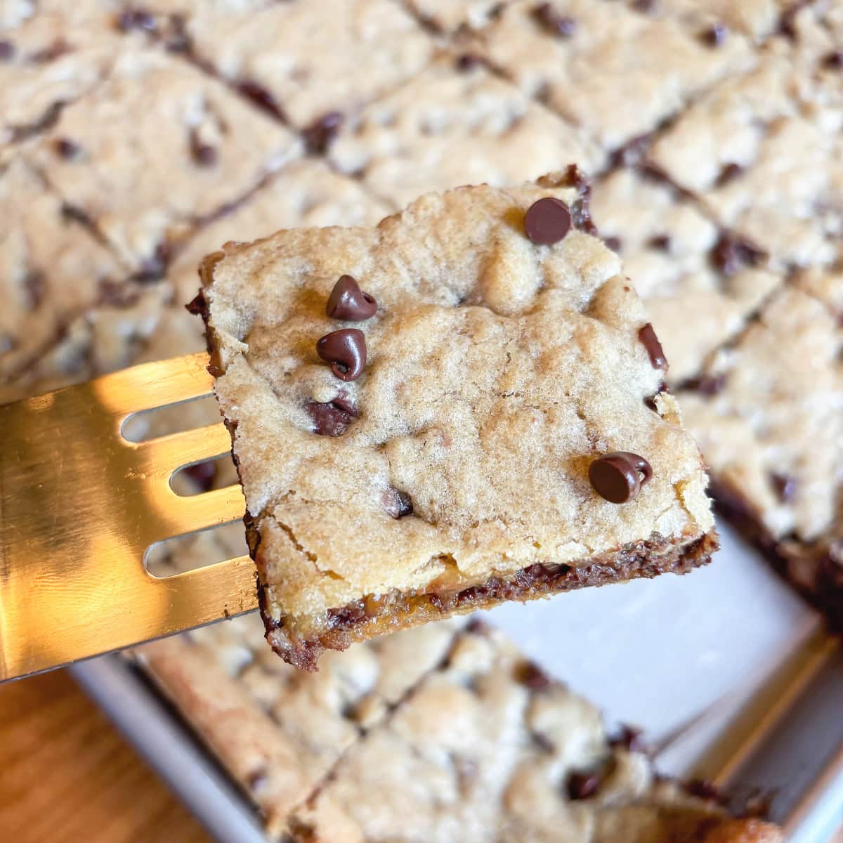 Chocolate chip cookie bar square lifted on a serving spatula above a pan of bars.