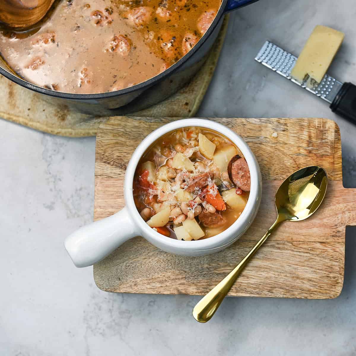 Cajun sausage and bean soup in a white bowl with a gold spoon beside it and soup pot in the background. Caption: A cozy serving of Cajun Sausage and Bean Soup with the pot nearby. Description: A wide shot showing a hearty bowl of Cajun soup filled with sausage, beans, and veggies, served with rice for the ultimate comfort food meal.