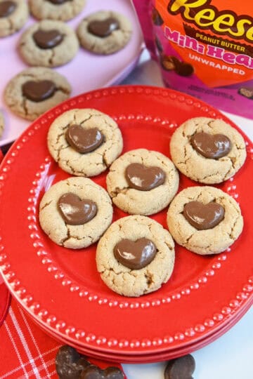 Peanut Butter Reese’s Heart Cookies stacked on a red plate with a bag of Reese’s heart candies in the background.