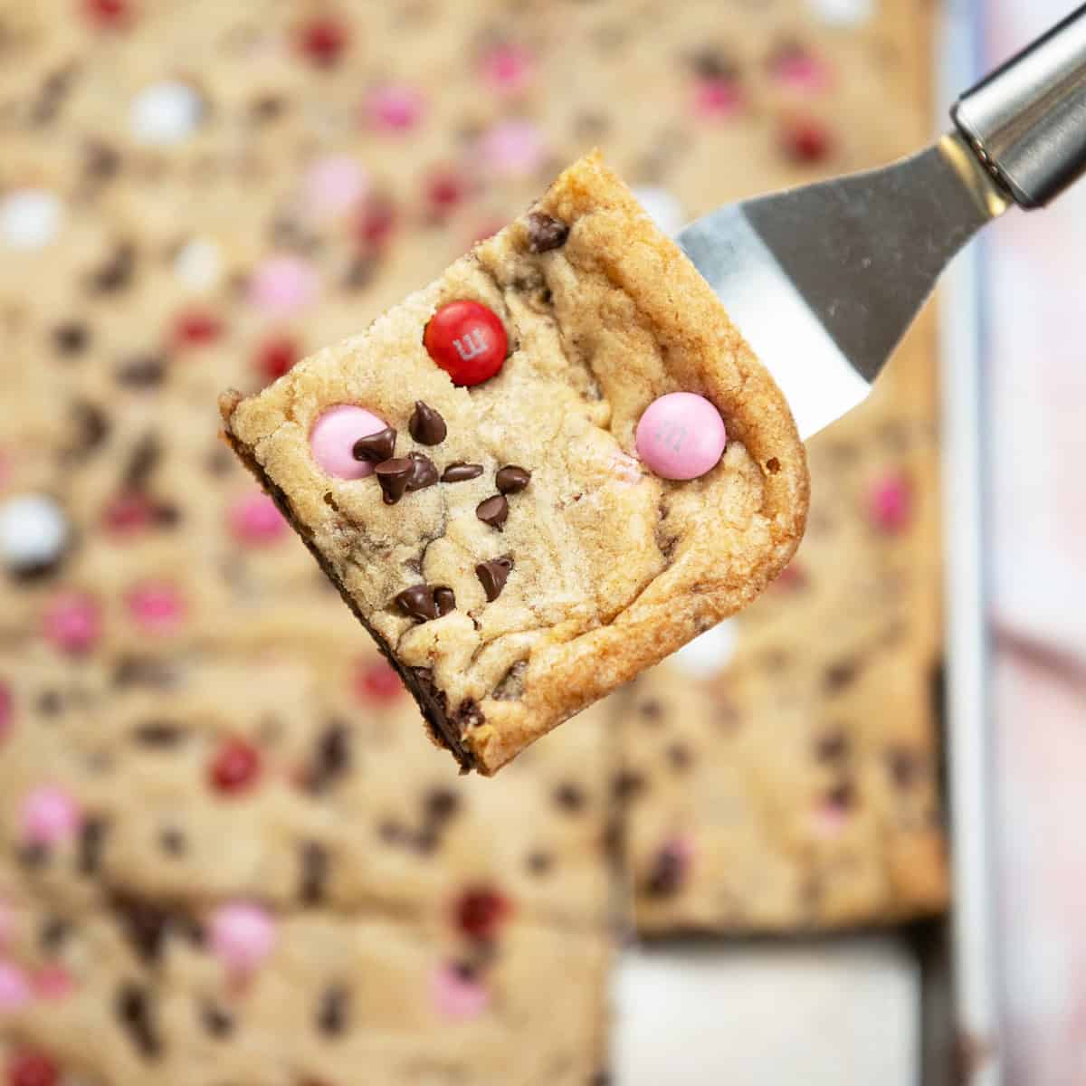 Valentine M&M Cookie Bar slice lifted above the baking pan showing thick, chewy layers and melty chocolate.
