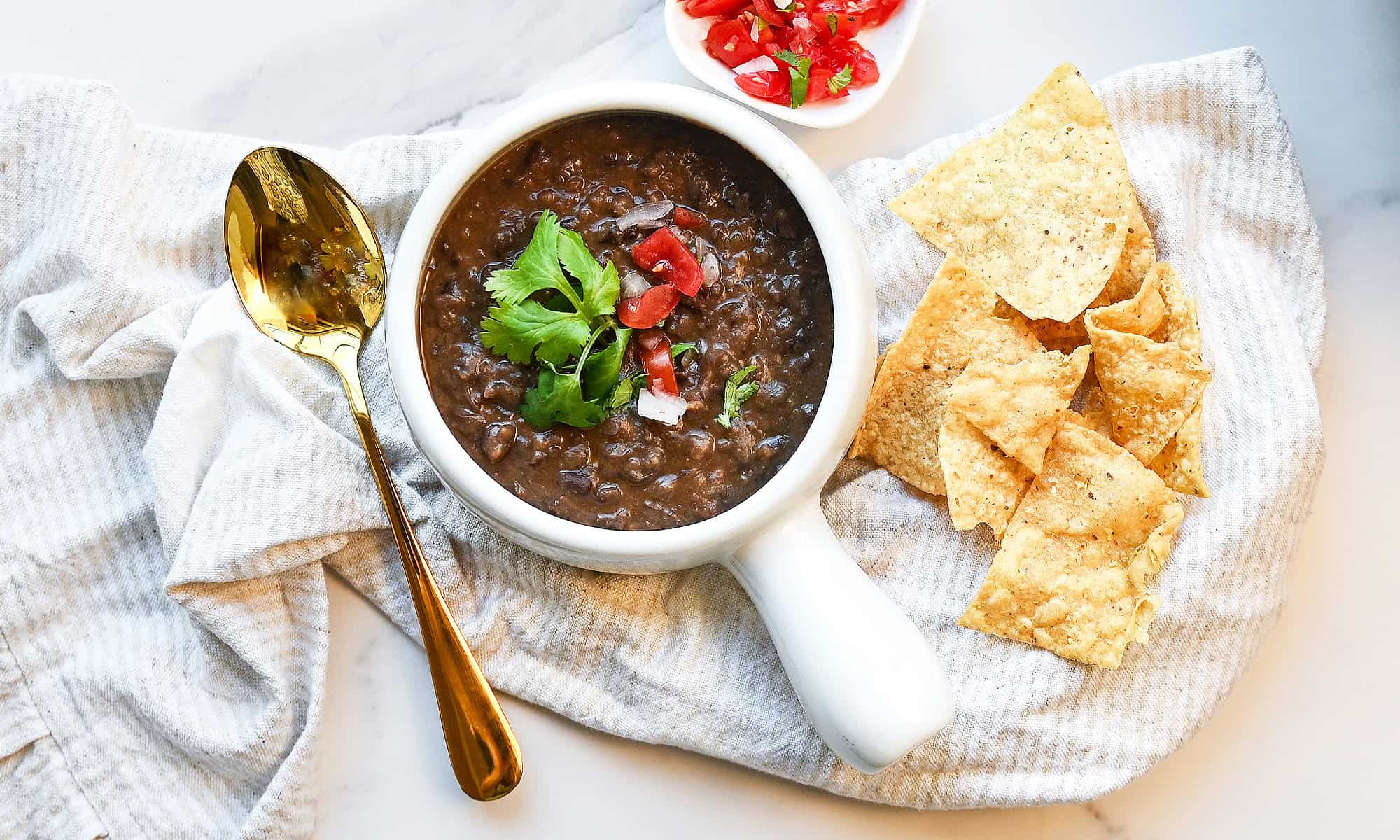 Close overhead view and round up size of thick black bean soup garnished with pico de gallo and cilantro in a white bowl with tortilla chips.