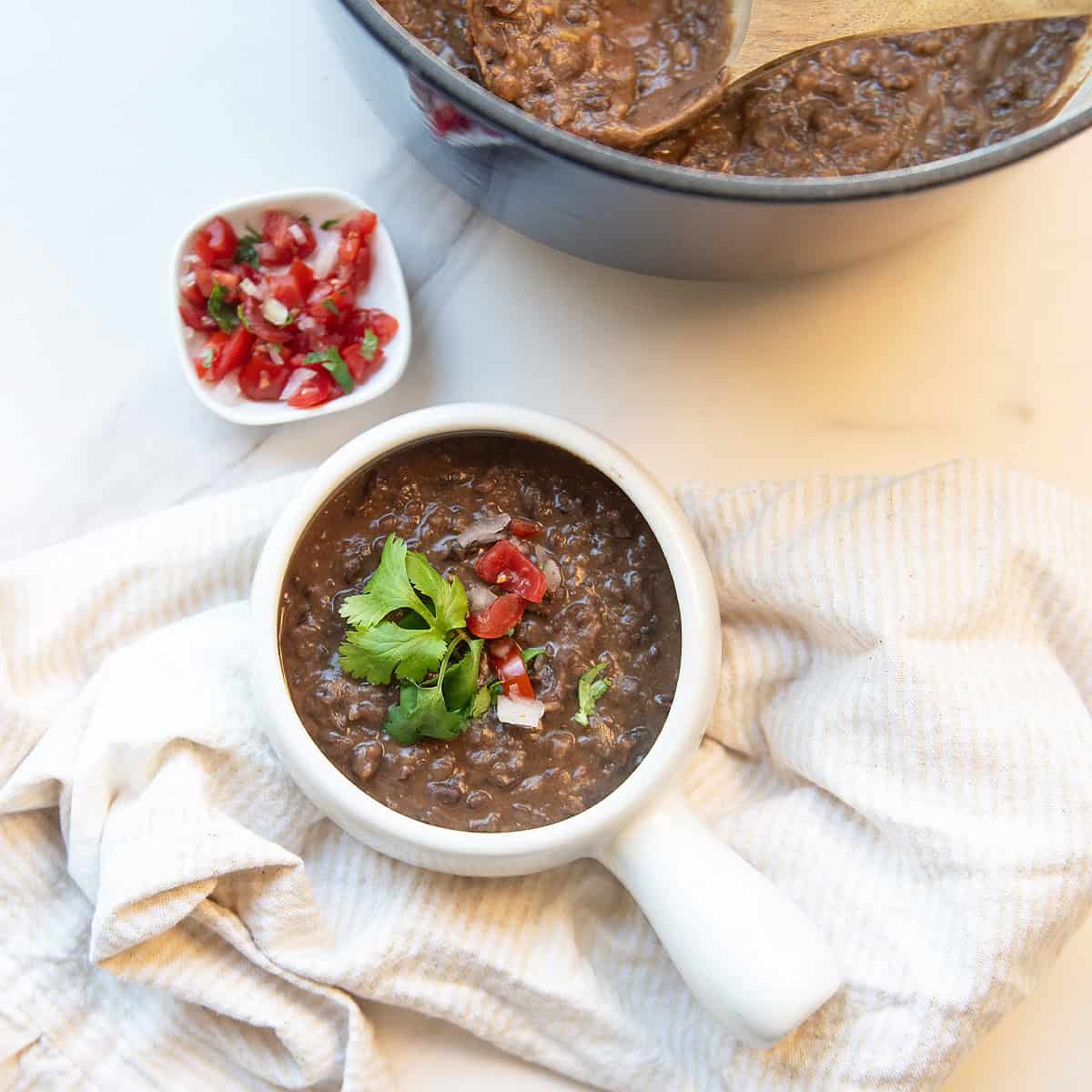 Overhead view of a white bowl of black bean soup garnished with pico de gallo and cilantro with a gold spoon and soup pot in the background.
