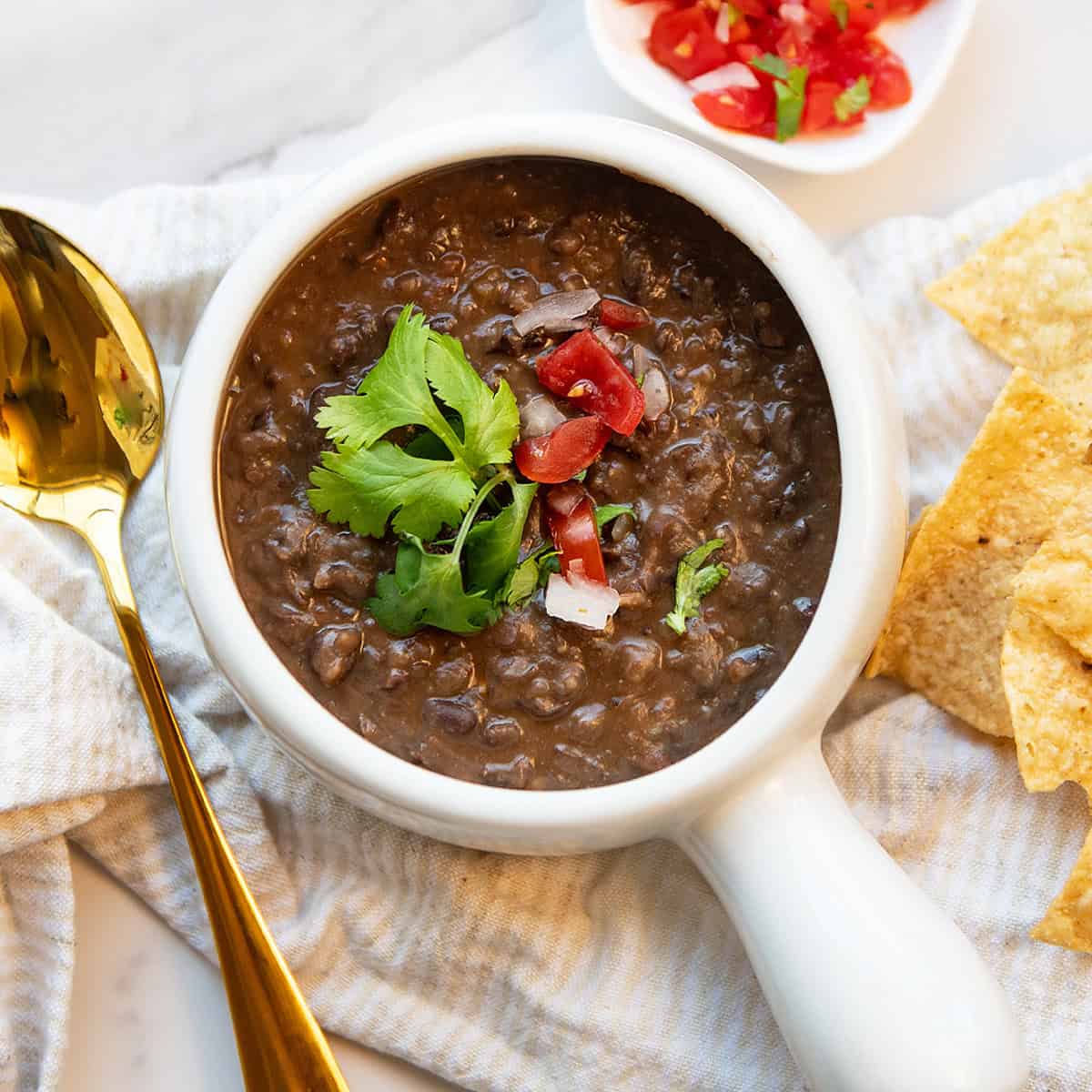 Close overhead view of thick black bean soup garnished with pico de gallo and cilantro in a white bowl with tortilla chips.