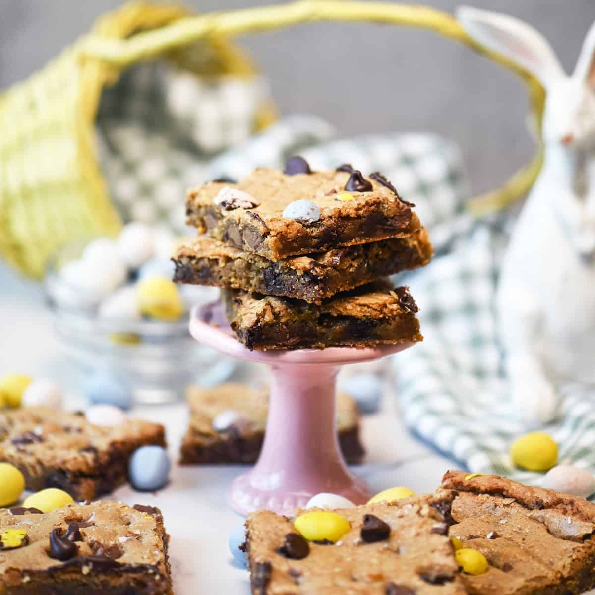 Three Cadbury Egg cookie bars stacked on a pink pedestal showing layers of chocolate chips and candy.