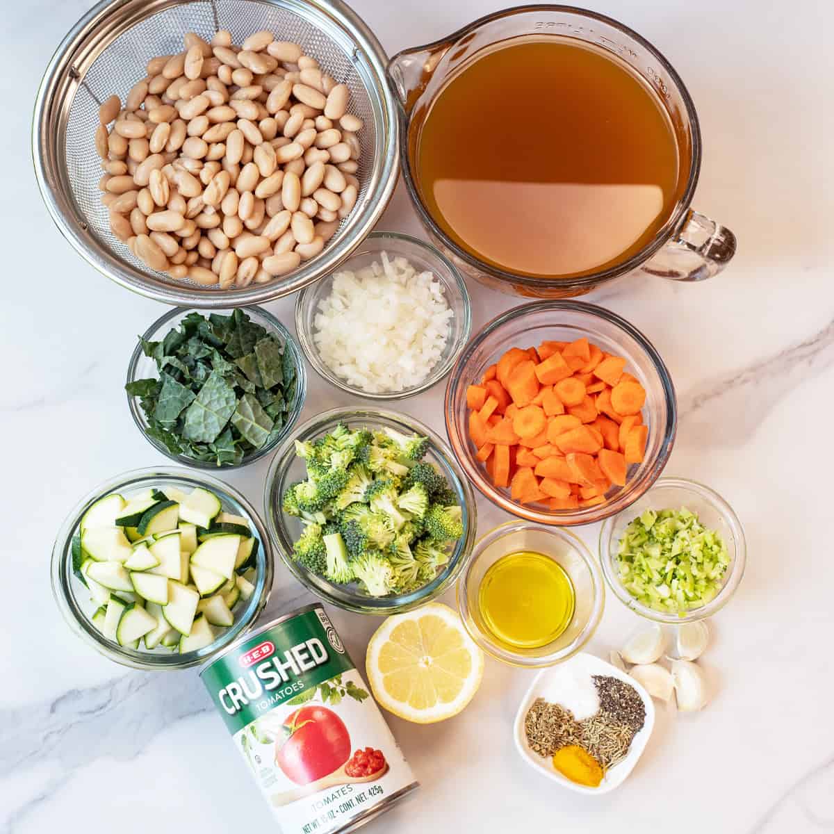 Ingredients for white bean vegetable soup including fresh vegetables, cannellini beans, broth, and seasonings arranged on a countertop.