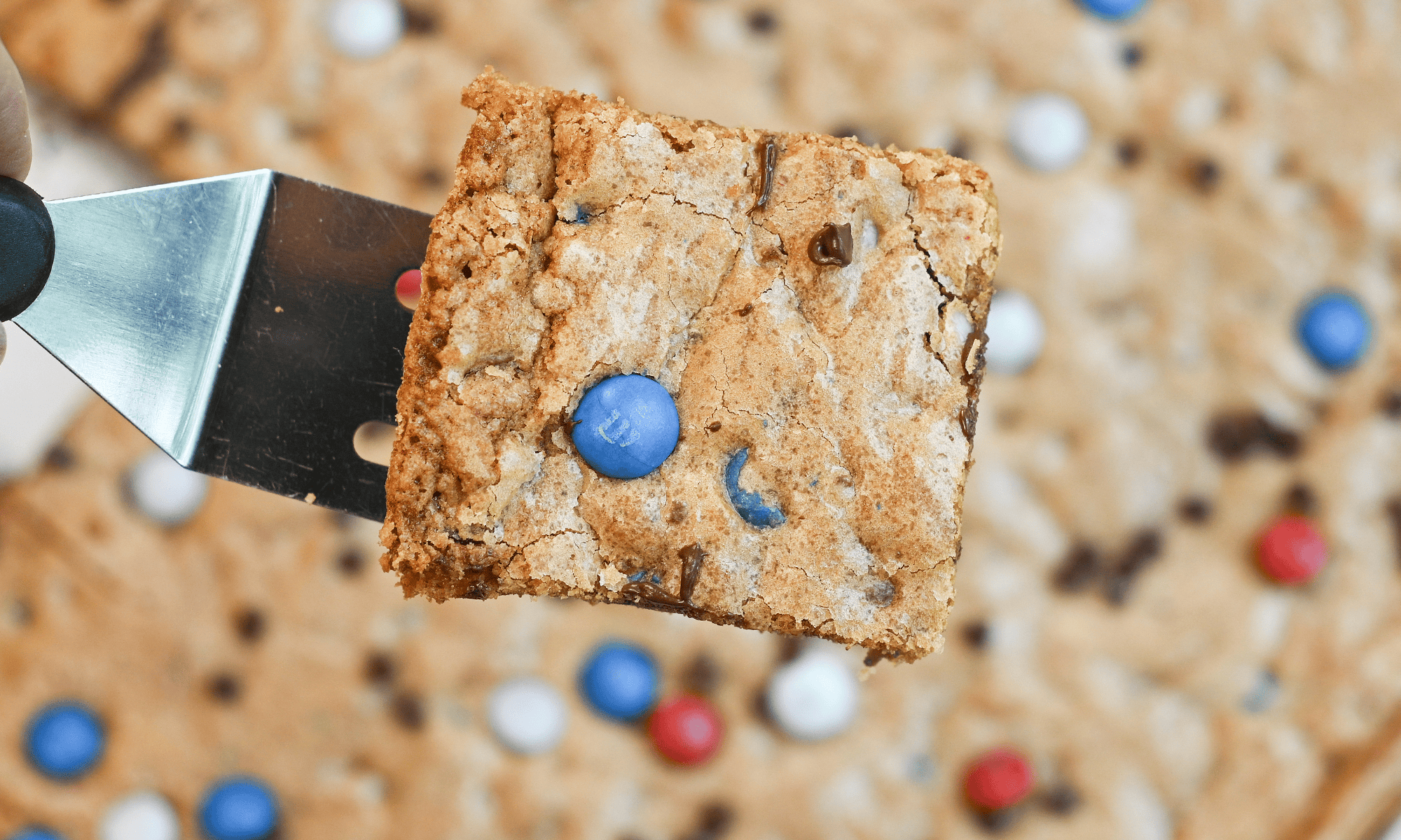 Overhead view of a patriotic M&M cookie bar lifted from the pan on a spatula with chocolate chips and colorful M&Ms.
