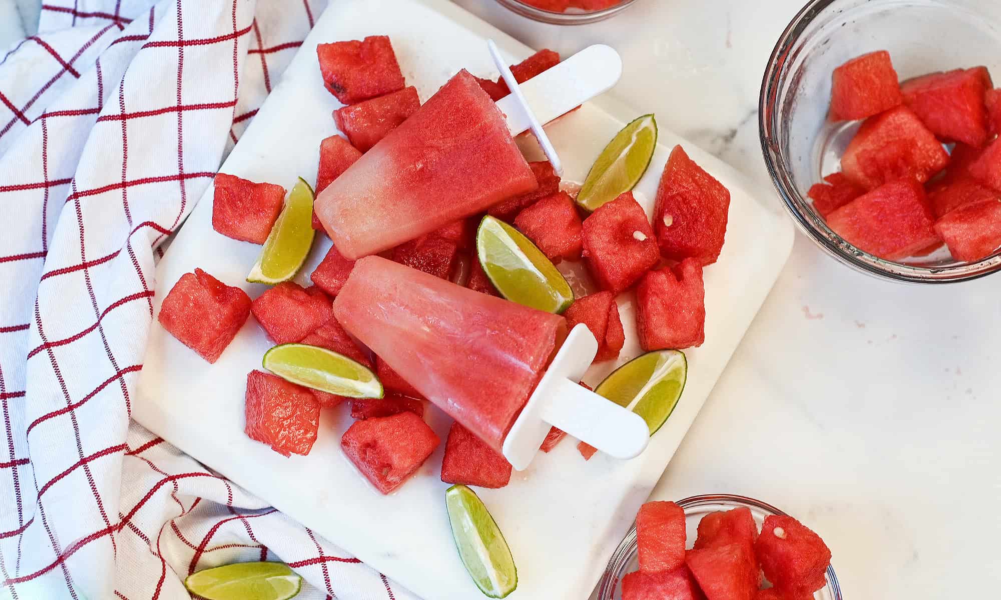 Two watermelon lime popsicles resting on a bed of fresh watermelon cubes and lime slices.