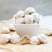 Snowball cookies coated in powdered sugar in a white bowl with one cookie cut in half showing the buttery pecan center.