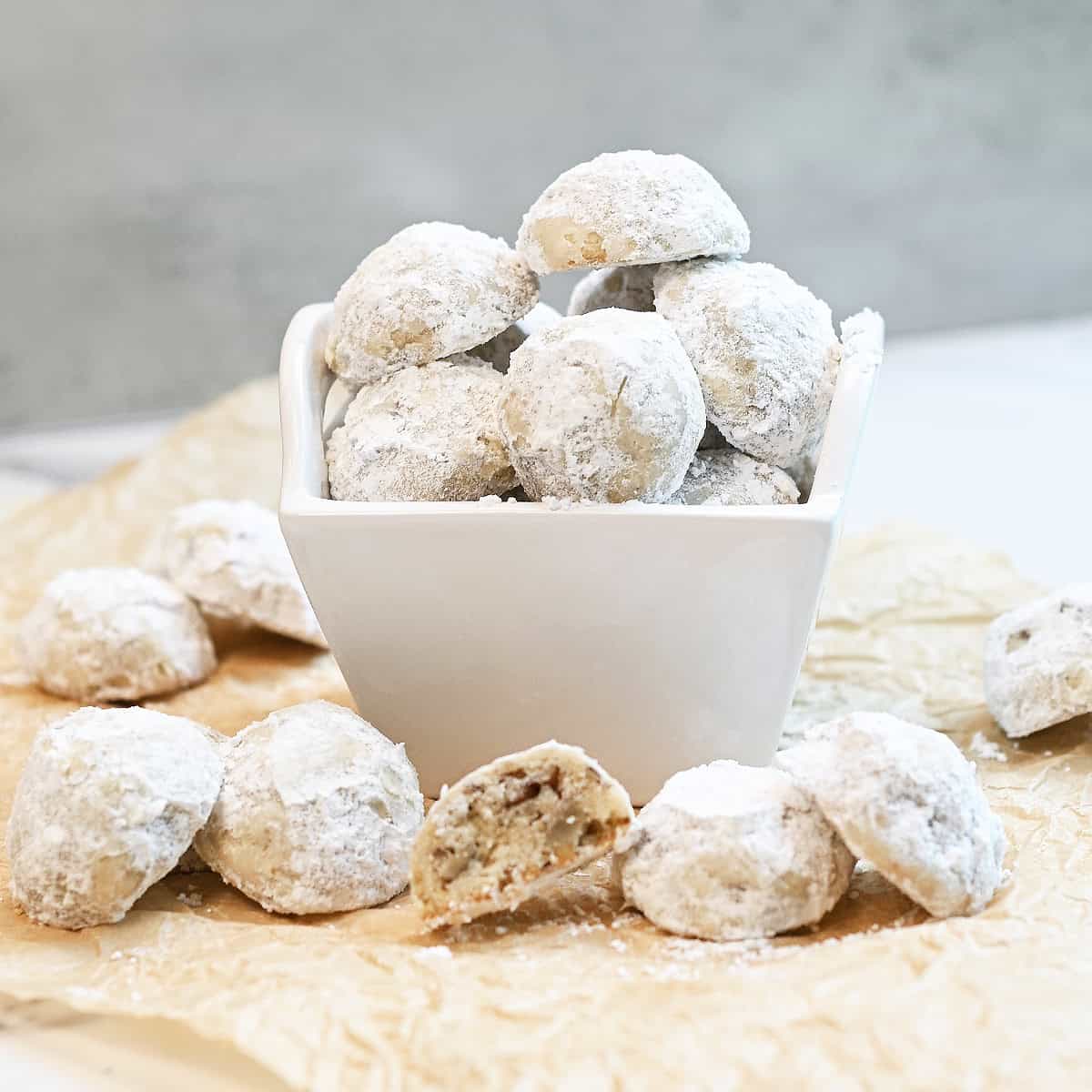 Snowball cookies coated in powdered sugar in a white bowl with one cookie cut in half showing the buttery pecan center.